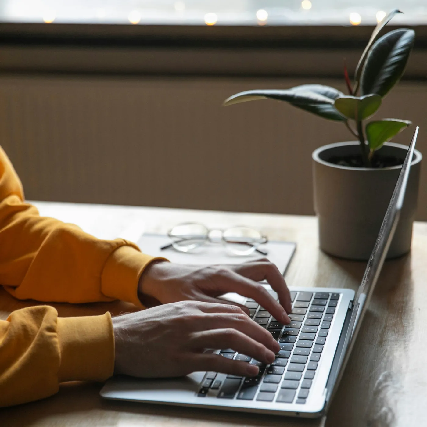 Hands typing on a laptop on a desk, with a pair of glasses and a pot plant beside it.